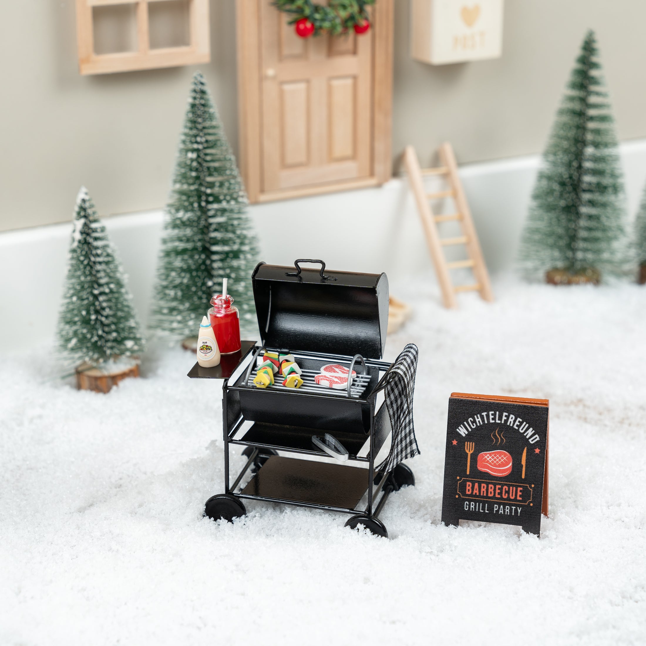 Wichtel-Barbecue-Set mit Miniatur-Grill, Saucen und Grillzubehör im verschneiten Wichteldorf vor einer festlich geschmückten Wichteltür.