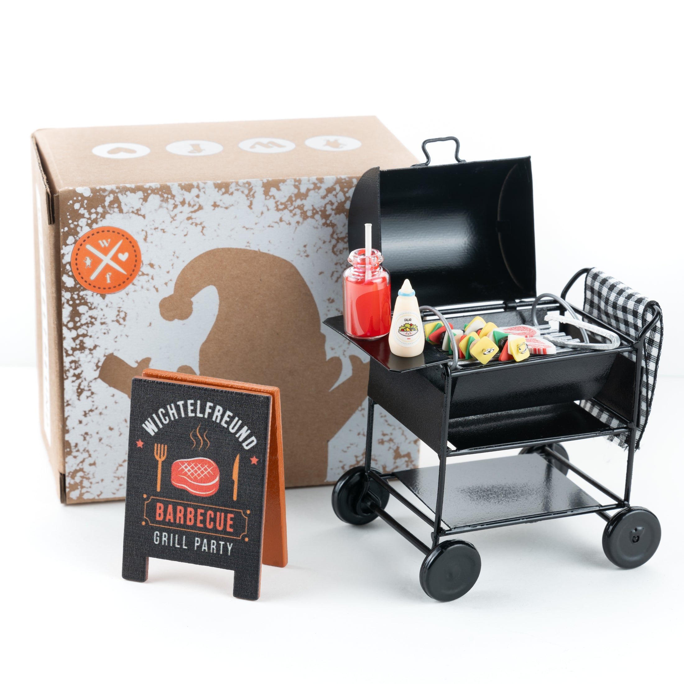 Wichtel-Barbecue-Set mit Miniatur-Holzkohlegrill, Grillgut, Saucenflaschen und „Wichtelfreund Barbecue Grill Party“-Schild vor dekorativer Verpackung.